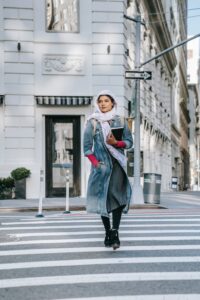 Full length confident lady in jean coat and headscarf with notebook in hand walking on crosswalk on city street at daytime and looking away